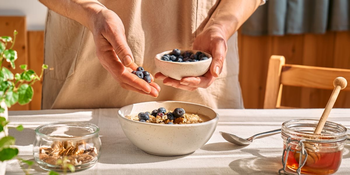 Female hand putting blueberries in the bowl with oatmeal porridge with walnuts and honey.
