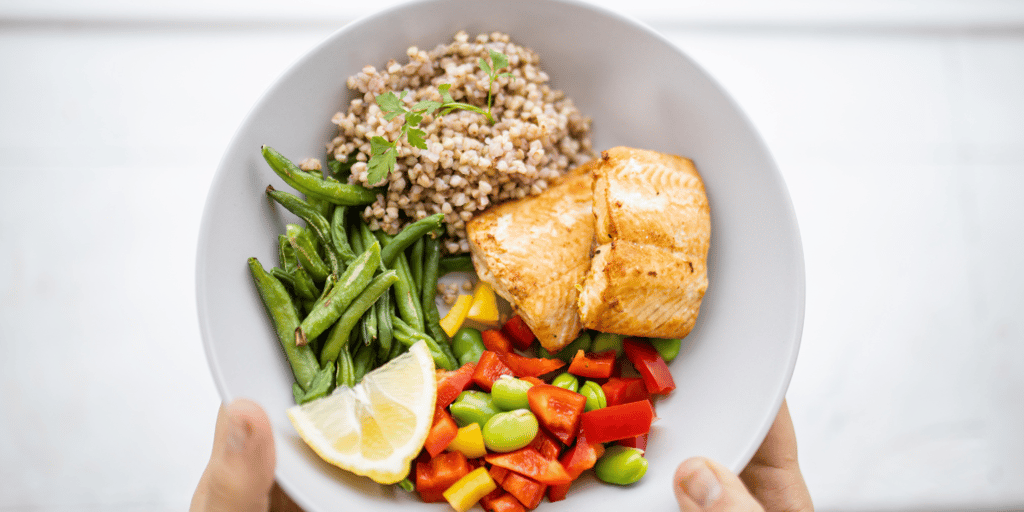 Hands holding salmon and buckwheat dish with green beans, broad beans, and tomato slices.