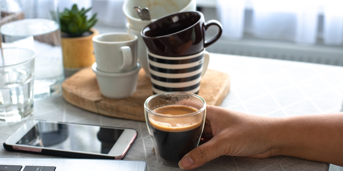 A hand holding a cup of espresso surrounded by empty cups of coffee, phone and laptop