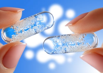 Fingers holding 2 semi-transparent pills containing probiotics and prebiotics