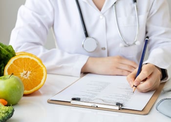 Female nutritionist writing diet plan on table with colorful fruits and vegetables
