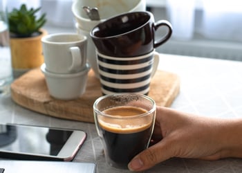 A hand holding a cup of espresso surrounded by empty cups of coffee, phone and laptop