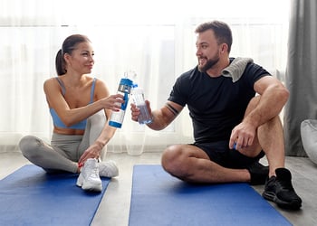 A man and a woman sitting on yoga mats in exercise attire drinking water